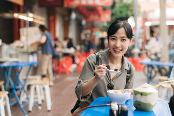 Happy young Asian woman backpack traveler enjoying street food at China town street food market in Bangkok, Thailand. Traveler checking out side streets.