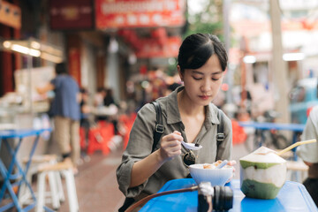 Happy young Asian woman backpack traveler enjoying street food at China town street food market in Bangkok, Thailand. Traveler checking out side streets.