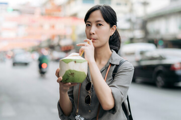 Happy young Asian woman backpack traveler drinking a coconut juice at China town street food market in Bangkok, Thailand. Traveler checking out side streets. 