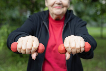 Smiling elderly woman is doing exercises with dumbbells outdoors in the park.  Focus is on hands. Active life of pensioners. Adaptation of pensioners in the modern world. 