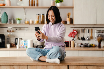 Cheerful asian woman using mobile phone while sitting on table in kitchen