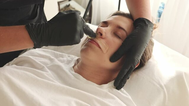 Waxing waxing specialist on a woman removing hair on her facial mustache using sugar paste. Woman is in pain during shugaring. Concept: beauty self-care depilation