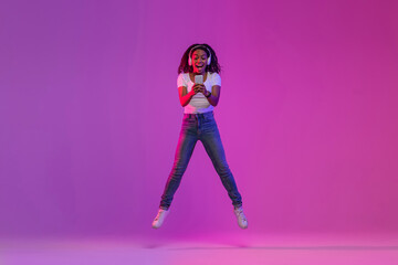 Joyful Black Woman With Smartphone In Hands Jumping Over Purple Gradient Background,