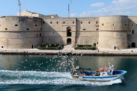 Aragonese Castle of Taranto with fishing boat returning to the port surrounded by dozens of seagulls. Puglia, Italy 