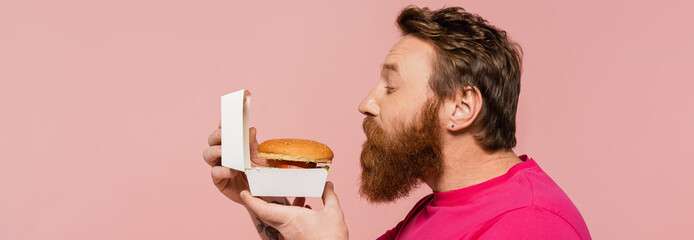 side view of bearded man smelling delicious hamburger isolated on pink, banner.