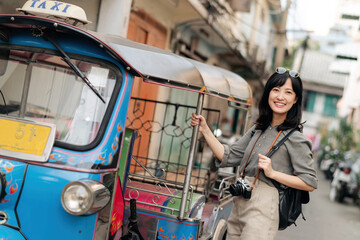 Fototapeta na wymiar Young Asian woman backpack traveler standing a side of Tuk Tuk taxi on summer vacations at Bangkok, Thailand. 