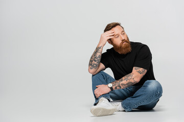 full length of thoughtful tattooed man in black t-shirt and jeans looking away while sitting on...