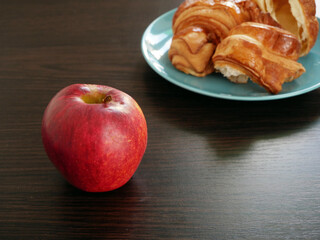 Naklejka na ściany i meble Red apple and croissants in plate on wooden background. Apple vs croissants, healthy food vs unhealthy food,