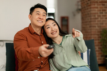 Happy korean spouses sitting on sofa in living room and watching TV together at home, free space