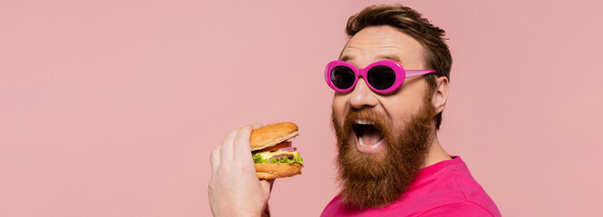 excited bearded man in stylish sunglasses opening mouth near hamburger isolated on pink, banner.