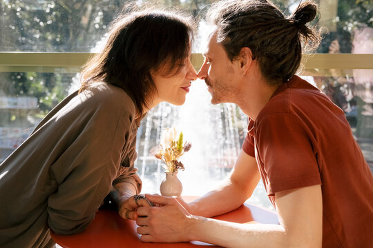 Young man and woman rub their noses and hold hands while sitting in a cafe on a warm sunny day.