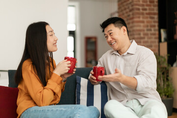 Loving korean couple drinking coffee and talking, enjoying free time together at home, sitting on sofa in living room