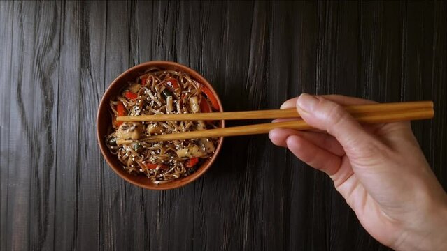 A woman's hand takes Chinese chopsticks soba noodles with chicken meat and vegetables from a clay bowl against a dark wooden table top view.