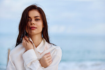 Portrait of a beautiful pensive woman with tanned skin in a white beach shirt with wet hair after swimming on the beach of the ocean sunset light with clouds, the concept of freedom and mental health 