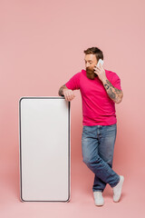 full length of bearded man in jeans talking on smartphone near white phone mock-up on pink background.
