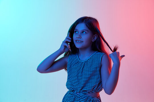 Little beautiful girl, child emotionally ralking on phone over gradient blue pink studio background in neon light. Concept of childhood, emotions, fun, fashion, lifestyle, facial expression