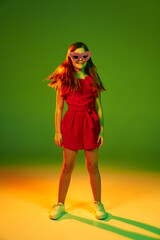 Little girl, child in red dress posing in furry sunglasses over gradient green yellow studio background in neon light. Concept of childhood, emotions, fun, fashion, lifestyle, facial expression