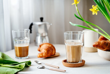 Spring cozy breakfast concept. Two glasses with coffee and milk, croissants, coffee pot and yellow daffodils. Morning, natural light