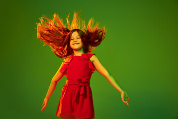 Little girl, child in red outfit posing with happy positive face over green studio background in neon light. Concept of childhood, emotions, fun, fashion, lifestyle, facial expression