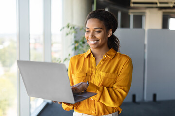 Successful african american businesswoman with laptop in hands standing near window in office interior, copy space