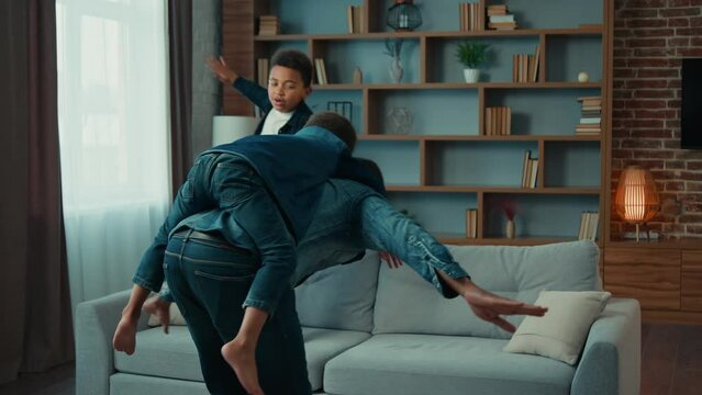 Happy African American father piggyback riding on back little preschool child son while sibling kid boy jumping on couch play airplane game. Dad with two boys children kids playing in cozy living room