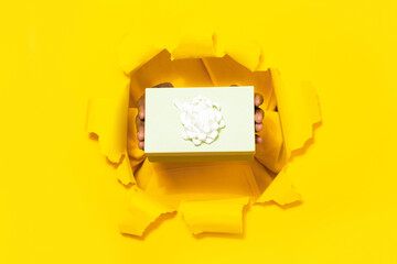Male hands giving wrapped gift box through torn yellow paper background, closeup, free space