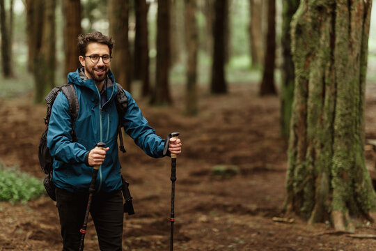 Smiling millennial caucasian guy in glasses and jacket with backpack and trekking sticks walking in cold forest