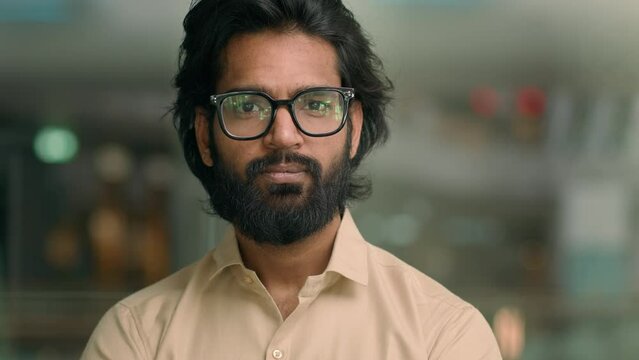 Medium Portrait Handsome Bearded Indian Entrepreneur In Glasses Standing In Creative Office Look To Side Turning Looking At Camera. Thoughtful Arabian Man Businessman Contemplating E-business Ideas