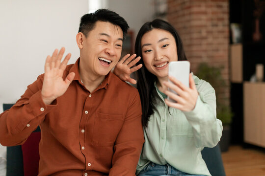 Technology, Meeting Remotely. Happy Asian Couple Having Video Call And Waving Hands At Cellphone Webcamera