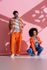 Interracial couple in sunglasses looking away on pink background with shadow.