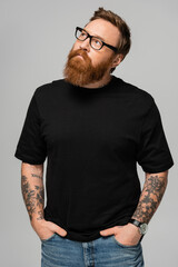 stylish bearded man in eyeglasses standing with hands in pockets of jeans and looking away isolated...