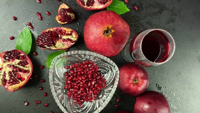 Table top view glass red wine or juice, ripe pomegranates, apples, green leaves on black background. Close-up fresh pomegranate arils. Cut open fruit scattered grains seed indoors. Studio moving shot
