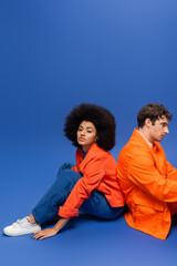 Curly african american woman in bright orange jacket sitting near man on blue background.