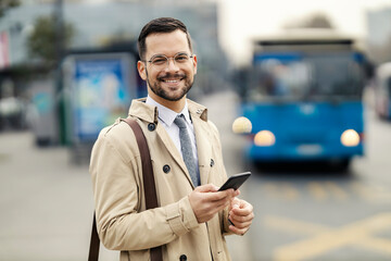 Portrait of a happy elegant man standing on a bus station and waiting for a public bus while using...