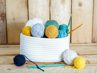 yellow, white and blue woolen balls in a white textile basket with crochet hooks on wooden ground - obrazy, fototapety, plakaty