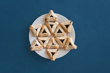 Happy Purim carnival decoration concept made from Hamantaschen or hamans ears cookies on the plate on blue background. Hebrew, jewish holiday celebration composition in shape of Star of David 
