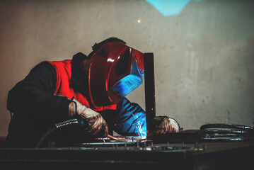 Profesional welder in protective uniform and mask welding metal pipe on the industrial table with other workers behind in the industrial workshop.