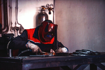 Profesional welder in protective uniform and mask welding metal pipe on the industrial table with other workers behind in the industrial workshop.