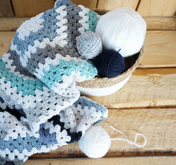 white, grey, blue granny square blanket with woolen balls in an white textile basket on wooden...