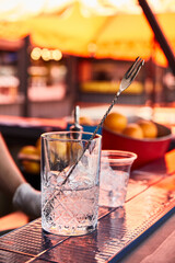a glass shaker with ice and a bar spoon, a glass for a summer cocktail with ice stands on a blue bar counter against the background of a summer bar with fruits, oranges, lemons, an orange umbrella on 