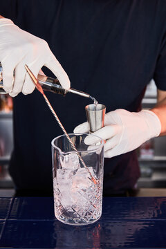 a bartender in white gloves and dark clothes prepares a cocktail, on a bar counter a measuring glass, glass shaker with ice and a bar spoon, a bartender pours alcohol into a measuring glass, jagger