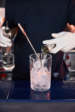 a bartender in white gloves and dark clothes prepares a cocktail, on a bar counter a measuring glass, glass shaker with ice and a bar spoon, a bartender pours alcohol into a measuring glass, jagger