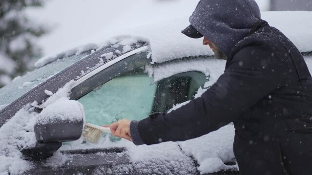 Man in hoodie cleaning snow-covered car with brush during heavy snowfall. Snowstorm, bad winter weather, natural disaster, slow motion. Snowstorm in winter season. Vehicle care in blizzard