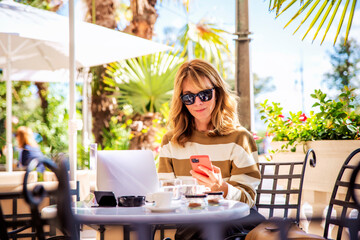 Happy woman with mobile phone and laptop sitting at outdoor cafe in summer