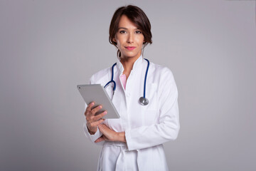 Studio portrait of middle aged female doctor using digital tablet for work and standing at isolated...