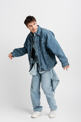 full length of stylish young man in denim jacket and jeans posing while looking away on grey.