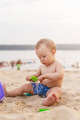 charming cute red-haired kid is sitting in the sand and playing with toys on the seashore