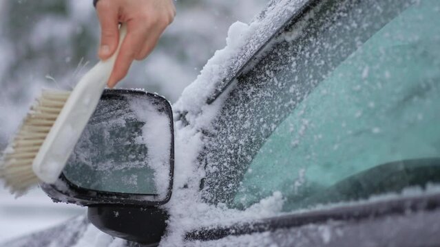 Close up cleaning snow from rear-view mirror of car with brush during snowfall cyclone slow motion. Snowstorm in winter season. Vehicle care in blizzard