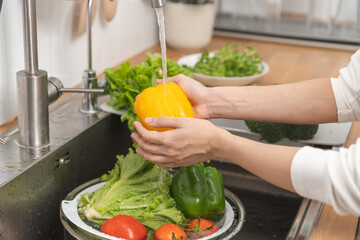 Attractive smile asian young housekeeper woman hand washing sweet pepper, yellow paprika, vegetables with splash water in basin of water on sink in kitchen, preparing fresh salad, cooking food meal.