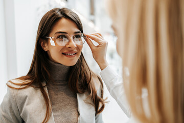 Beautiful and fashionable woman choosing eyeglasses frame in modern optical store. Female seller...